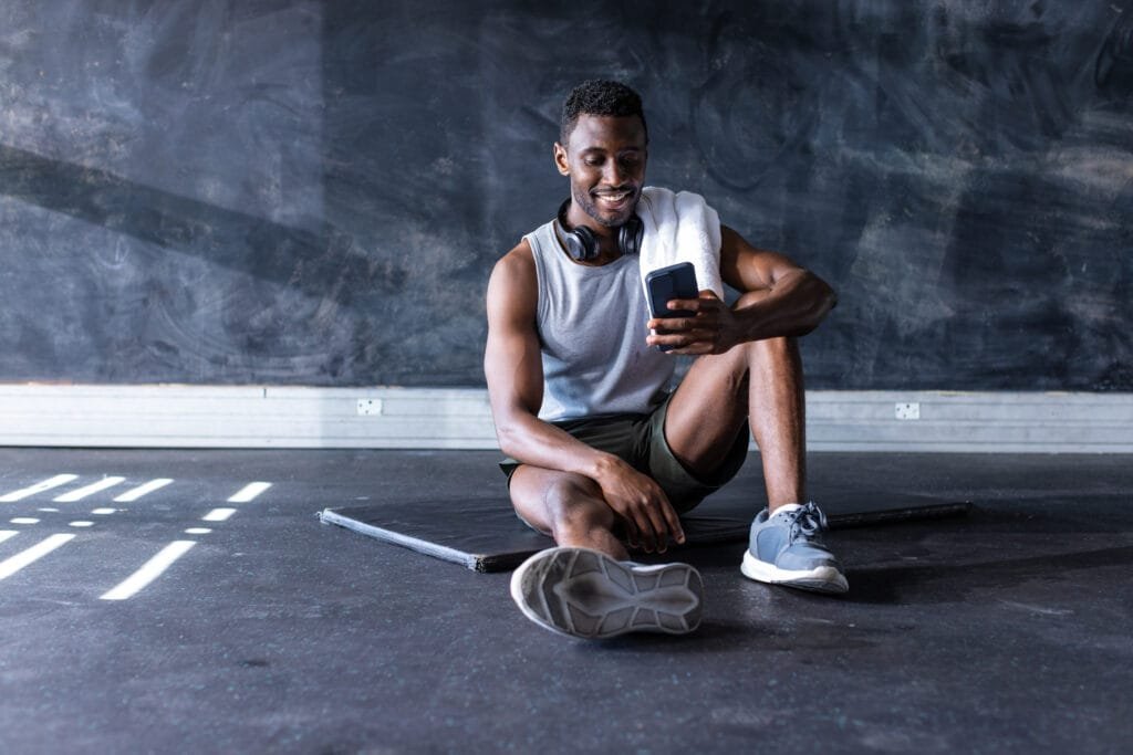 African American young male athlete sitting on floor in gym, looking at smartphone, copy space. He's wearing casual gym clothes and has headphones around his neck, unaltered.